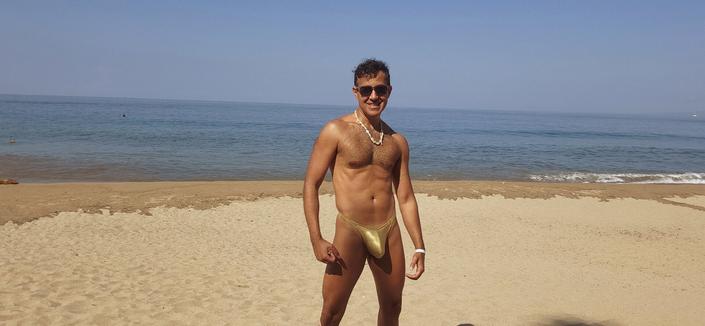 Selfie, standing on a sandy beach w/ the ocean behind, wearing a shiny swim thong.