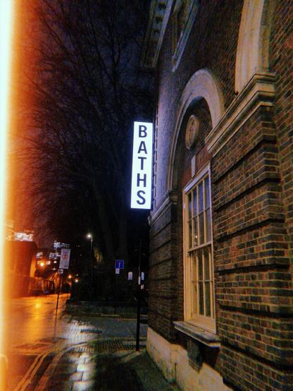 A vertical lit up white sign on the side of a building reads BATHS 
In the background is a dark street with pavement shiny with rain.