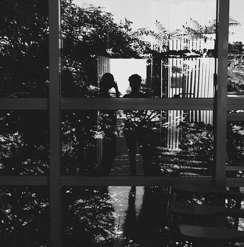 A black and white photograph shows the silhouettes of two people reflected in a large, multi-paned window. The figures appear to be in conversation, though their expressions are obscured. The reflection is layered over a view of lush foliage and a modern building exterior, creating a complex interplay of foreground and background. The image has a mysterious and somewhat melancholic tone.