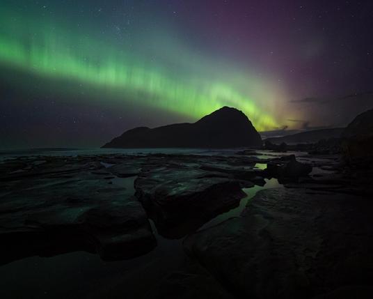 Stunning view of Lion Rock in Tasmania under the vibrant green and purple hues of the aurora australis, with rocky terrain and reflective water pools in the foreground under a starry night sky.