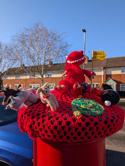 A crocheted letterbox topper on a traditional red British postbox. It's a bright day with clear blue sky and there is a terrace of houses in the background and a car parked nearby. The postbox topper is red and is decorated with further works of knitting craft in the form of animals associated with the Chinese zodiac. A horse, monkey, rabbit, and chicken can be seen, and there is a coiled snake at the front with a toothy, smiling dragon sat in the centre on the top holding a stick with a yellow flag on which is written "Kung Hei Fat Choy".