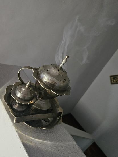 On a small metal tray, there's a metal fluted container with a cover and handle with smoke coming out of the holes on top. Next to it is a smaller container in the same design but no handle. There's a pair of tongs next to them, and a small perforated metal dish. There's no other ornamentation on the metal.