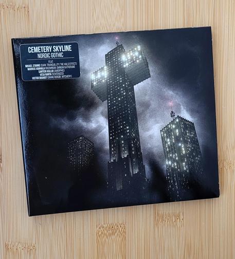 A CD album laying on orange-brown wood. CD cover is dark, almost black, but three high rise buildings with a lot of lit up windows are in the middle, partially wrapped by clouds. The central building has the shape of a cross, with brighter lights on the arms of the cross. Artist name Cemetery Skyline and album name "Nordic Gothic" are on a sticker in the upper left corner, with smaller text naming the band members and their other bands: Mikael Stanne (Dark Tranquillity, The Halo Effect), Markus Vanhala (Insomnium, Omnium Gatherum), Santeri Kallio (Amorphis), Vesa Ranta (Sentenced), Victor Brandt (Dimmu Borgir, Witchery)