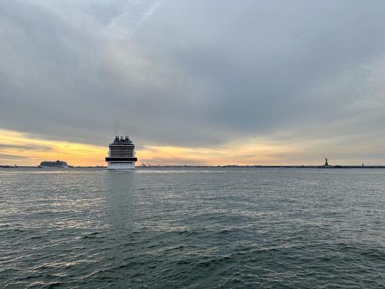 Photo looking across New York Harbor on a cloudy approaching sunset. The ass-end of a cruise-ship is just to the left, and to the left is another cruise-ship in profile sailing off to left (i.e., to the Narrows). Over to the right is the Statue of Liberty.