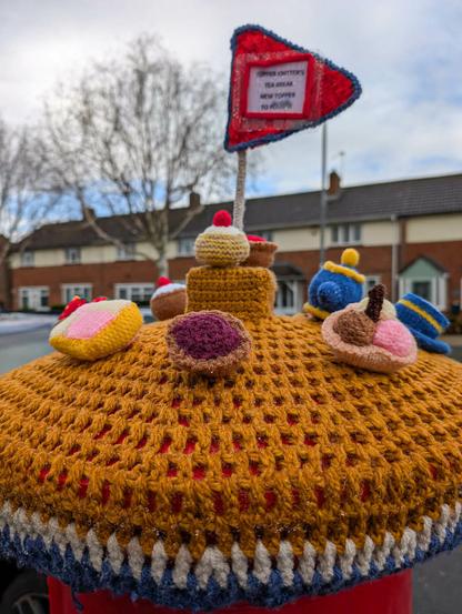 A crocheted postbox topper on a postbox. There are houses in the background, out of focus, and the sky is mostly cloudy but with a few blue patches showing. The topper is primarily an orange colour and has knitted cakes on the side we can see plus a small teapot, cup, and saucer on the rear. A red flag is raised in the centre on which the words "Topper Knitters Tea Break. New Topper To Follow" can just be made out.