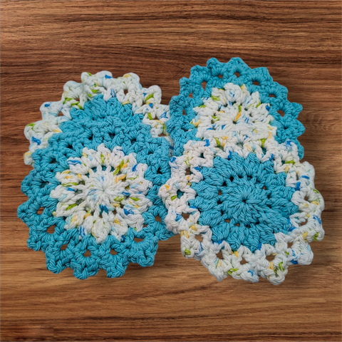 A set of four crocheted coasters in turquoise and a white, yellow, green, and blue multicolor yarn. The pattern looks floral and two coasters are multi in the center and turquoise on the outside while the other two are the opposite.