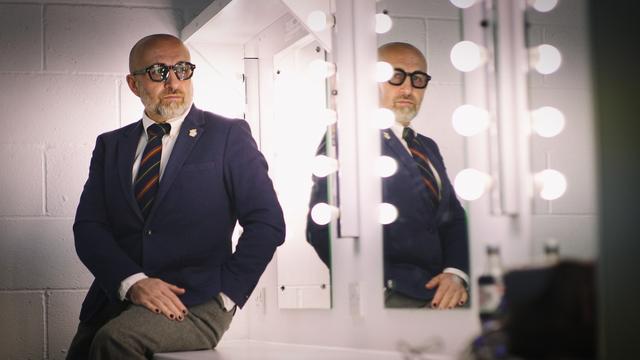 me, a white middle aged guy, wearing cool thick framed glasses, a blue blazer, white shirt, blue patterened tie and grey trousers, sitting on my dressing table backstage, reflected in the mirror.