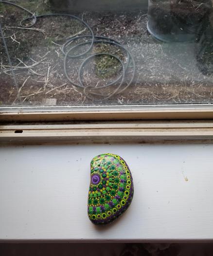 A human ear shaped rock on a white windowsill. The ground outside can be seen through the window. The rock is painted with a variety of green and purple dots in a semicircle starting with a large purple dot on the flat side of the rock (on the left in the picture) and then rows of smaller dots around it from there, working to the right. The entire top of the rock is covered in the dots.