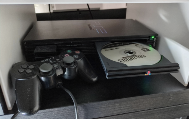 A running Ps2 Fat with an opened disk tray showing the game disk of Final Fantasy X International next to a PS2 controller.