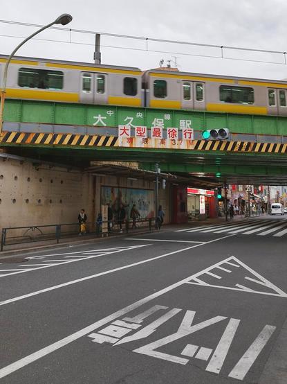 Portrait shot of a road and the railway bridge over it. A JR commuter train, silver with bright yellow livery, is passing over it so a small section of that train made the shot. The bridge is made of massive concrete pillars and a steel truss put on top, painted turquoise. The name of the station is written on the side as well as a height restriction warning for the traffic underneath. The road is wet with rain. There are plenty of white markings on the road including one for a bus stop. Through the opening of the bridge we can see a bit of the shopping street that extends from the bridge.