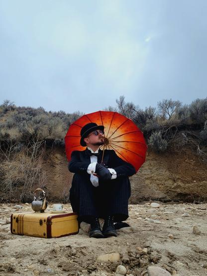 In my best antique French tuxedo and bowler hat, I sit on a rock having afternoon tea. My red and cream parasol rests on my shoulder, giving me a halo akin to a poppy flower. Next to me is a vintage Samsonite suitcase where a teapot and teacup rest. A makeshift table when civilization is far away. I'm sitting on a dried lake bed where the old shore edge, covered in dried brush, can be seen behind me. Gazing up at the sky, I dream of picnics and fêtes with friends, but it's a solitary existence when far from civilization and breaks for tea are only a momentary reprieve.
