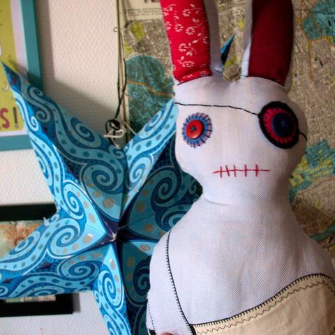 Light blue hand-made stuffed bunny with felt eye patch like a pirate and a sewn on button for the other eye and a chainsaw made out of of white fabric in her paw. Behind her a cyan-coloured star shape out of paper and framed prints and maps.