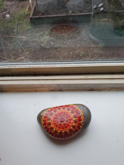 A small, oblong rock on a white windowsill. An orange, yellow and red painted circle is in the center, made up of small dots of paint.
