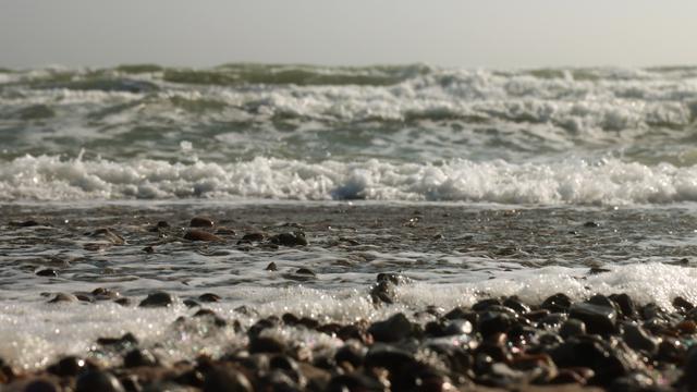 Auflaufende hohe Wellen auf einen Strand mit rundgeschliffenen Steinen. Zwischen den Steinen glitzert der Schaum des Meeres.