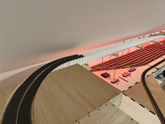 A model railway baseboard in the corner of a room. A crossing and a set of points leads to a double track that turns across the back of the room whilst descending.