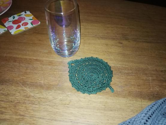 Donkergroen haakwerkje in de vorm van een blaadje.
Het is bijna een rondje, bestaande uit meerdere ringen, maar doordat de ringen aan één kant dikker zijn, loopt het rondje dasr uit in een puntje. Aan andere kant zit een steeltje. 
Het blaadje ligt op een houten tafelblad. Ernaast staat een glas.
Het werkje is ca 10 cm in doorsnede.