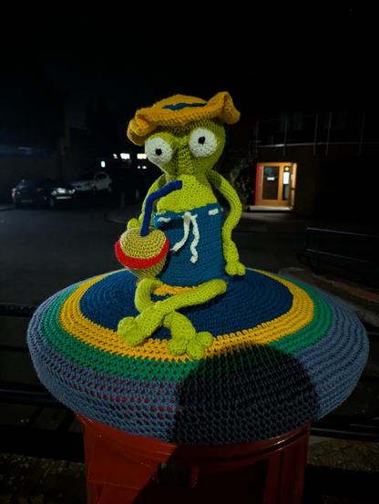 A colorful crocheted figure resembling a green, cartoonish frog wearing a yellow sun hat and blue swimwear, holding a drink with a straw. The figure is positioned on a vibrant, multi-colored crocheted seat atop a red mailbox, it is nighttime.