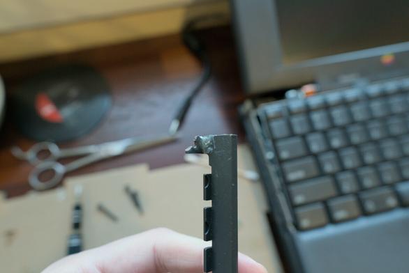 Photo of a particular plastic component of the PowerBook (the left hinge cover which wraps around the left hand side of the keyboard) being held up with the rest of the PowerBook showing in the blurry background. The part has a plastic lip sticking out that used to have a through-hole for as screw. This through-hole part has broken in half, but I epoxy-glued back the remaining half so that this piece can hold on to the screw at least a little bit still. It is not strictly a load bearing piece, fortunately, otherwise this would have not done any good.