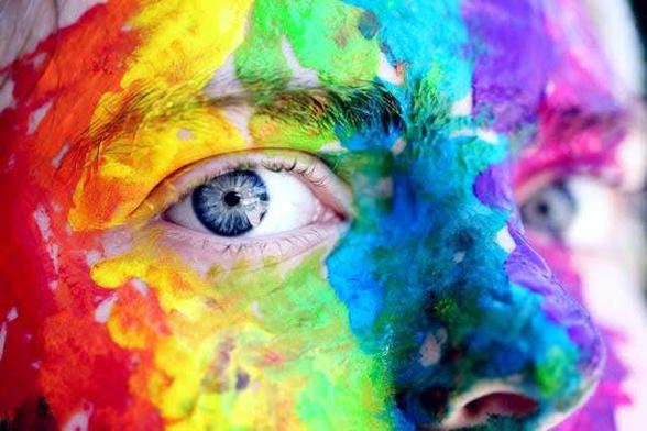 Primer plano de un ojo y parte de la cara de una persona cubiertos con pintura de colores vibrantes. Los colores incluyen rojo, naranja, amarillo, verde, azul y morado, aplicados con un estilo abstracto y salpicado. El ojo es azul y está parcialmente oculto por la pintura, pero es visible y se centra en la imagen. La pintura da la impresión de una explosión de color sobre la piel
