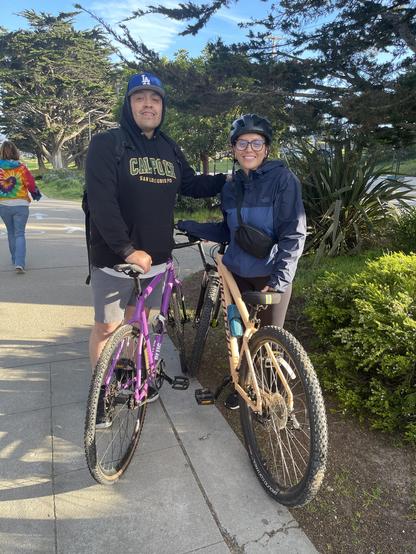 A smiling duo—a father and his young adult daughter—stand alongside their bicycles on a paved, car-free multiuse trail.