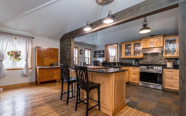 A picture of the traditional style kitchen in our potential new home.  The ceiling is vaulted and follows the roofline.  The flooring in the small breakfast area is thin strip oak, and the 12 by 12 tiles in the kitchen are different shades of brown.  There are two elevated chairs at a centre island.  All of the cabinetry is a honey colour pine with a black countertop and stone looking backslash.