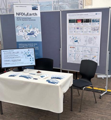 NFDI4Earth booth at FOSSGIS: a table with flyers and a computer screen in front of a banner and an academic poster