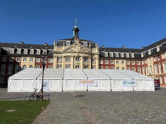 A large tent in front of the Münster Castle with banners for the FOSSGIS conference and OpenStreetMap and OSGeo