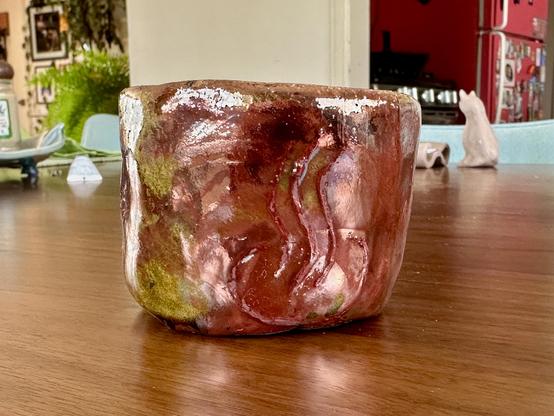 A small handmade ceramic cup that has been raku fired to a very coppery finish with spots of golden yellow. The emoji symbol for hot spring is etched on the front and filled with a red underglaze (the copper mixed with it a little).