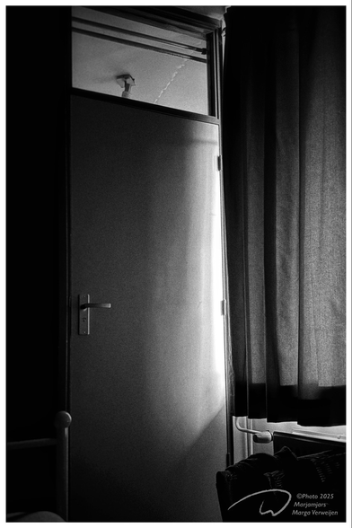 In the right corner of the bedroom, a door in the wall is lit by the penetrating daylight that comes in from behind and between the curtains. A bedpost can be seen in the still dark opposite corner. under the curtain, the outline of a chair in front of the heater.