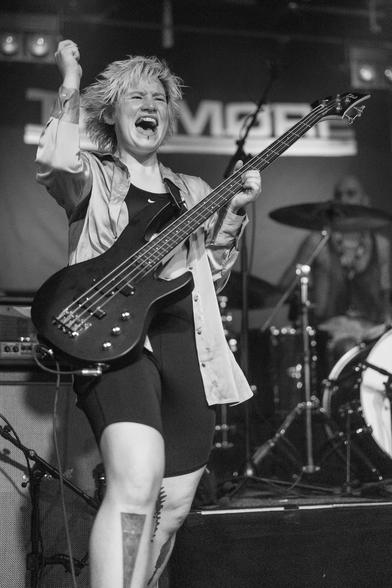 A musician passionately performing on stage, playing a black bass guitar. She has short, spiky blonde hair and is wearing a black outfit with a loose shirt. The background features drums and stage lighting, creating a vibrant concert atmosphere.