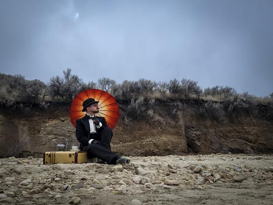 I'm pausing for a solitary tea party while in the middle of nowhere. Everything is dry and the sky is awash in grey. Sitting on a rock in my best French tuxedo and bowler I look out over the dried lake bed I have found myself in. A vintage Samsonite suitcase rests on the ground next to me functioning as a makeshift table to hold my teapot and teacup. I rest my red and orange parasol on my shoulder which creates a glowing halo effect behind me.