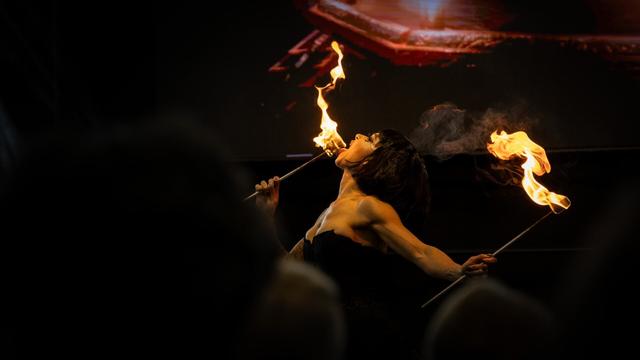A fire performer dressed in black engages in a fire-eating act, holding two flaming torches. She tilts her head back, bringing one of the flames close to her face, touching the torch with her tongue. The other torch burns in the air. The dimly lit background and audience silhouettes frame the dramatic moment.