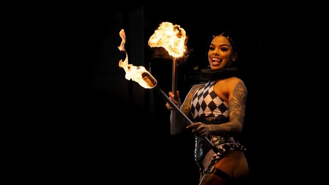 A fire performer dressed in a black-and-white harlequin-style outfit holds two flaming torches. She has an expressive face, sticking her tongue out playfully, and her tattoos are visible. The background is dark, making the firelight stand out dramatically.