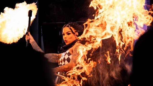 A fire performer in a harlequin outfit waves a large flaming torch, creating a massive burst of fire in the foreground. Her expression is focused, and her tattoos and braided hairstyle are visible. The fire engulfs much of the frame, creating a visually striking effect.