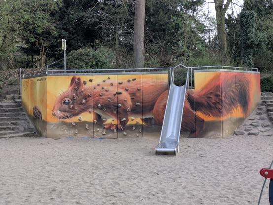 Photo of a playground. In the middle of the photo is a wall, about 2 meters high, limited by stairs in the left and right. On the middle to right side of the wall is a slide and some boulder stones for climbing middle to the left. The wall is painted with a realistic graffito of a giant red squirrel pointing its nose to a walnut.