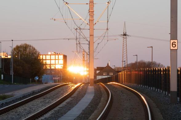 The view follows a double-track light rail line to the horizon. There, the rising sun is reflected in the glass façade of a high-rise office building so that it looks as if it is about to explode in an orange fireball. The light is reflected on the running surfaces of the rails