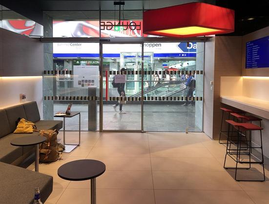 A view of the lounge: grey and red furniture, a glass back wall opening a view to the main hall with people passing by. My luggage and shoes lying around in an untidy manner.