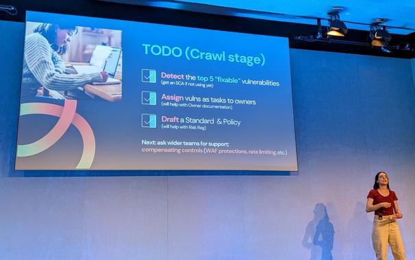 Celine Pypaert stands on stage at QCon London in front of a presentation slide titled 'TODO (Crawl stage)'. The slide lists three completed security tasks, each with a checkmark: 'Detect the top 5 "fixable" vulnerabilities', 'Assign vulns as tasks to owners', and 'Draft a Standard & Policy'. Each task includes a note about its benefit, such as helping with documentation or risk registration. The slide ends with a note: 'Next: ask wider teams for support; compensating controls (WAF protections, rate limiting, etc.)'. A photo of a woman working on a laptop is shown on the left side of the slide.
