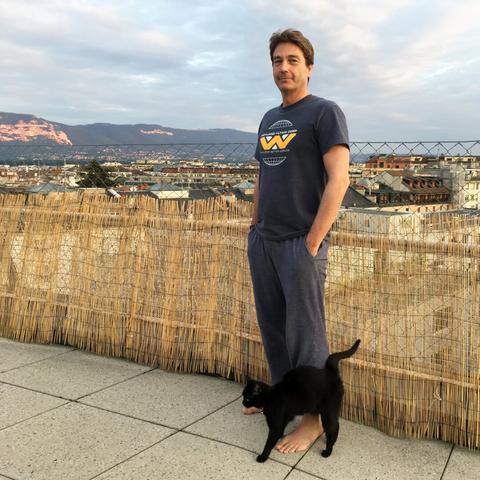 A black cat and yours truly, the latter wearing a “Building Better Worlds” Weyland-Yutani tee, standing on a terrace with a view over Geneva and the mountains.