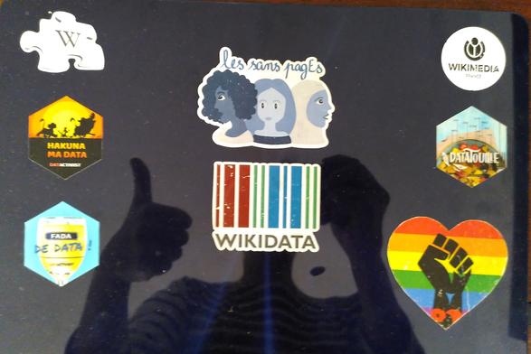 Ombre d'autoportait avec pouce levé sur ordinateur avec stickers Wikimedia France, Wikidata, Les Sans Pages, autour de la donnée (Datactiviste) et un coeur arc-en-ciel avec un poing levé