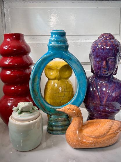 A colourful group of ceramic items including an orange duck, a purple buddha sculpture, a yellow owl, a red vase, a blue round vase with a see through area, and a green porcelain vase with a frog sculpture on top.