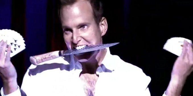 Gob from Arrested Development doing a magic show with cards fanned in both hands and a knife in his mouth, with a deranged look on his face.