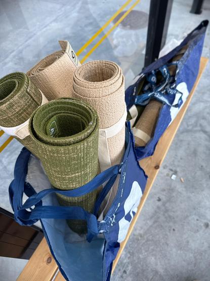 A blue reusable shopping bag sits on a wooden bench, filled with rolled-up yoga mats in earthy tones—two green and two beige. Another similar bag, partially visible, holds additional mats and a dark strap or harness. The scene appears to be at an outdoor or semi-covered space with a concrete floor and yellow lines, possibly at a bus stop or transportation hub.