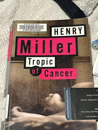 a paperback book, featuring a sepia-toned photograph of a lady reclining topless, taken from the belly up. there is a bookmark covering their chest. white and pink boxes indicate that this is “tropic of cancer” by henry miller.