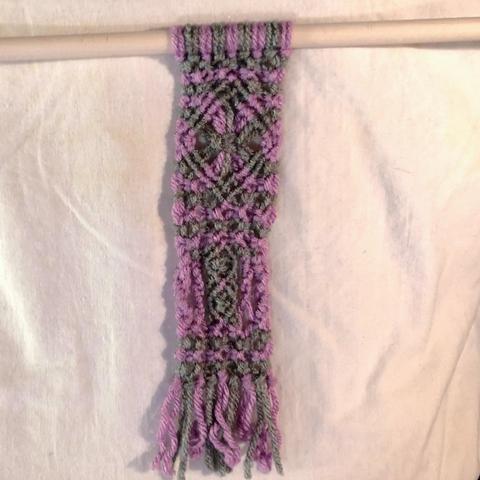 A macrame piece in lavender and gray on a dowel. There is a diamond motif with what looks like as starburst at the center on top and two small ovals on top of each other flanked by twisty rope like bits below and then, of course a fringe at the bottom