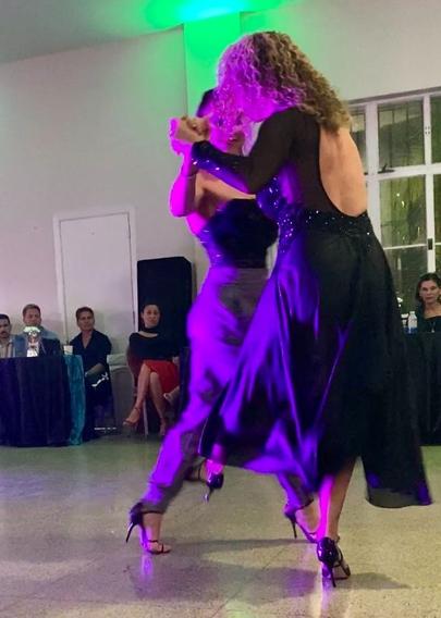 Liliana, with blonde curly hair and a long open-backed gown kicks between the legs of Martina, who is in grey pants and a lacy strapless top, as she takes a forward step.