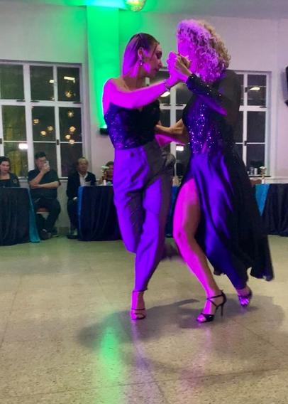 Liliana, with blonde curly hair and a long open-backed gown leads Martina, who is in grey pants and a lacy strapless top. They’re in Americano position, Liliana standing on her right leg and Martina on her left leg, torsos toward each other, with the other (outside) legs extended behind them.