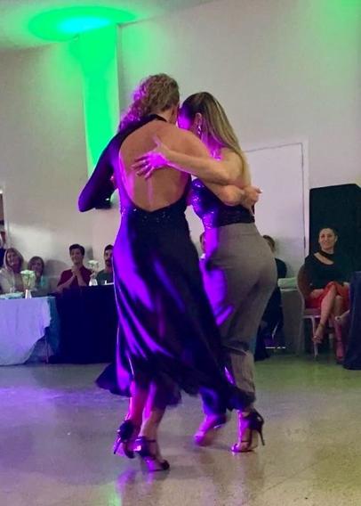 Liliana, with blonde curly hair and a long open-backed gown leads Martina, who is in grey pants and a lacy strapless top. They are taking tiny steps with their knees bent as they dance to fast music, and Martina is rotated, moving around Liliana counter clockwise.