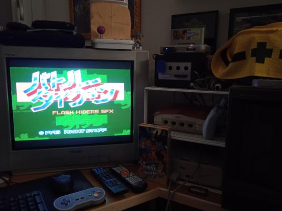 Una postazione di retrogaming con una TV a tubo catodico accesa, con una schermata verde e delle scritte giapponesi. A destra ci sono diverse vecchie console impilate una sull'altra.