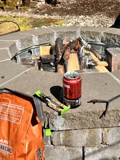 A fire pit with logs and burning flames, a can of imperial cherry cider, a savory jalapeño nut bar, and a bag of charcoal are arranged on a stone surface. The setting includes a natural background with moss and rocks. Probably a backyard someone spent a lot of time building during COVID but neglects now.